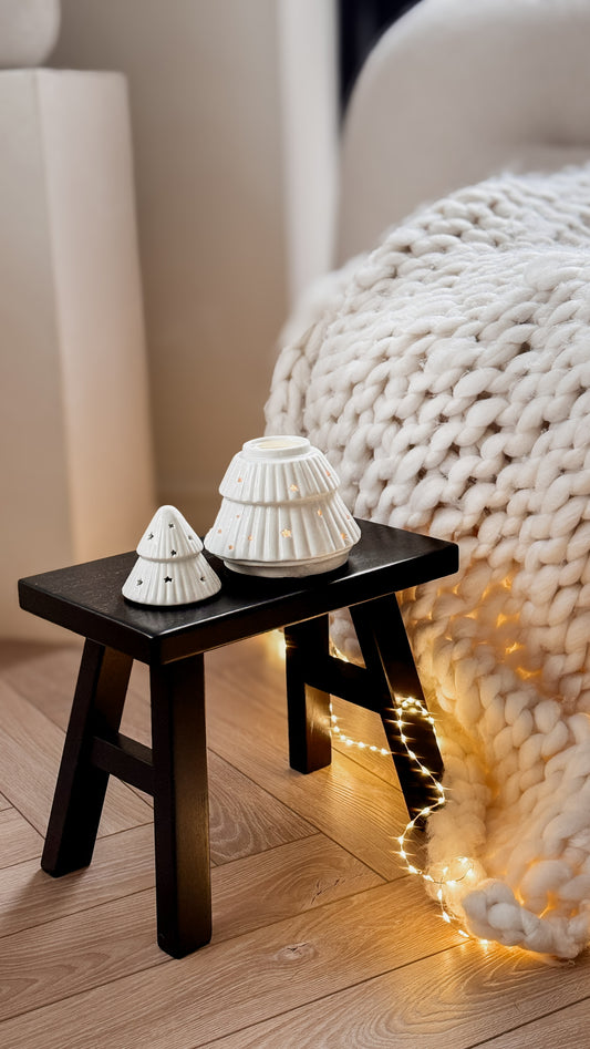 Small black wooden stool with white decorative christmas tree wax melt burner on a wooden floor next to a textured white blanket.