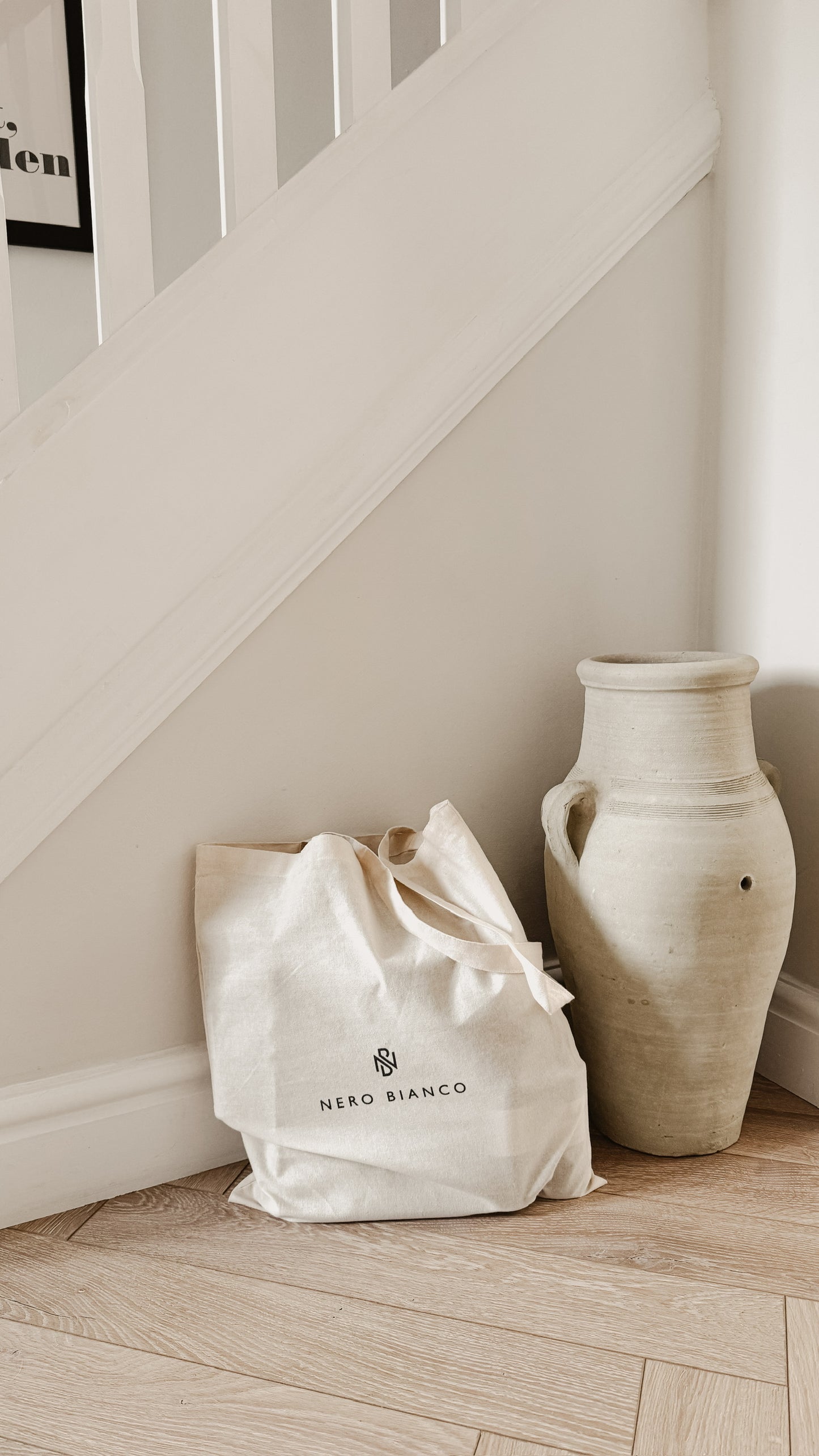 Nero Bianco tote bag and ceramic vase on a wooden floor against a white wall.