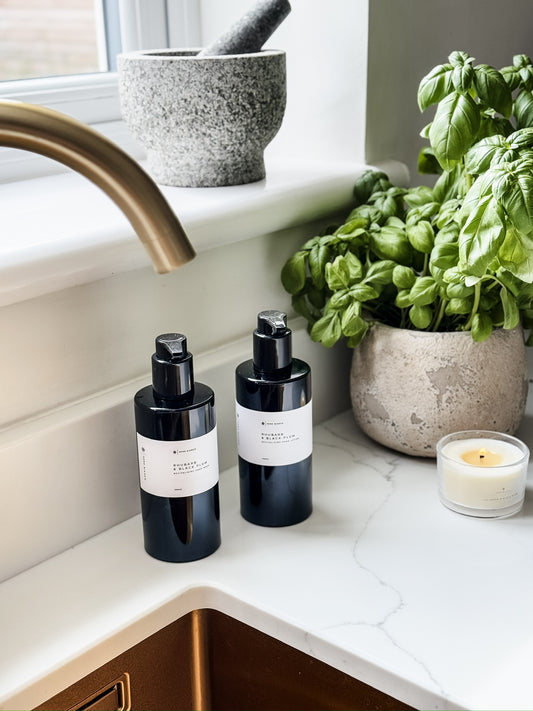 hand wash and hand lotion in black bottles with white labels on a kitchen counter next to a basil plant and candle.