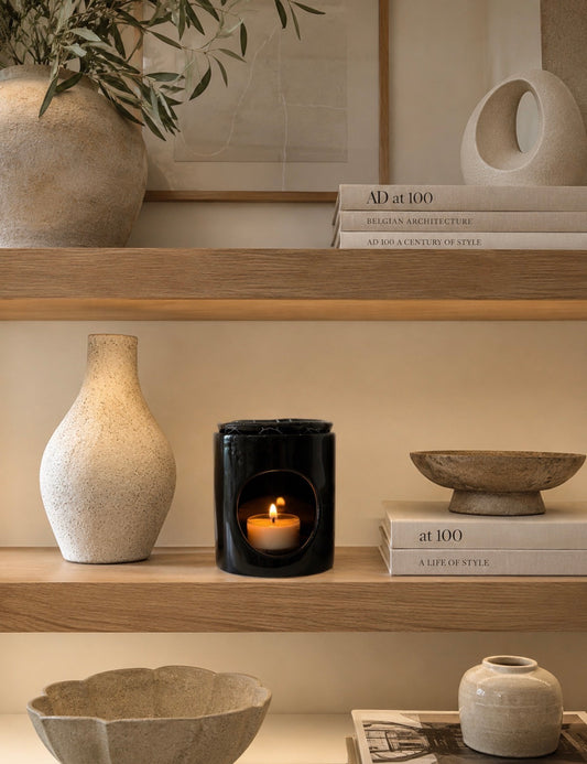 A black ceramic candle holder with a lit candle inside, styled on a wooden shelf alongside beige ceramic vases, bowls, and books in a warm, minimalist interior setting.