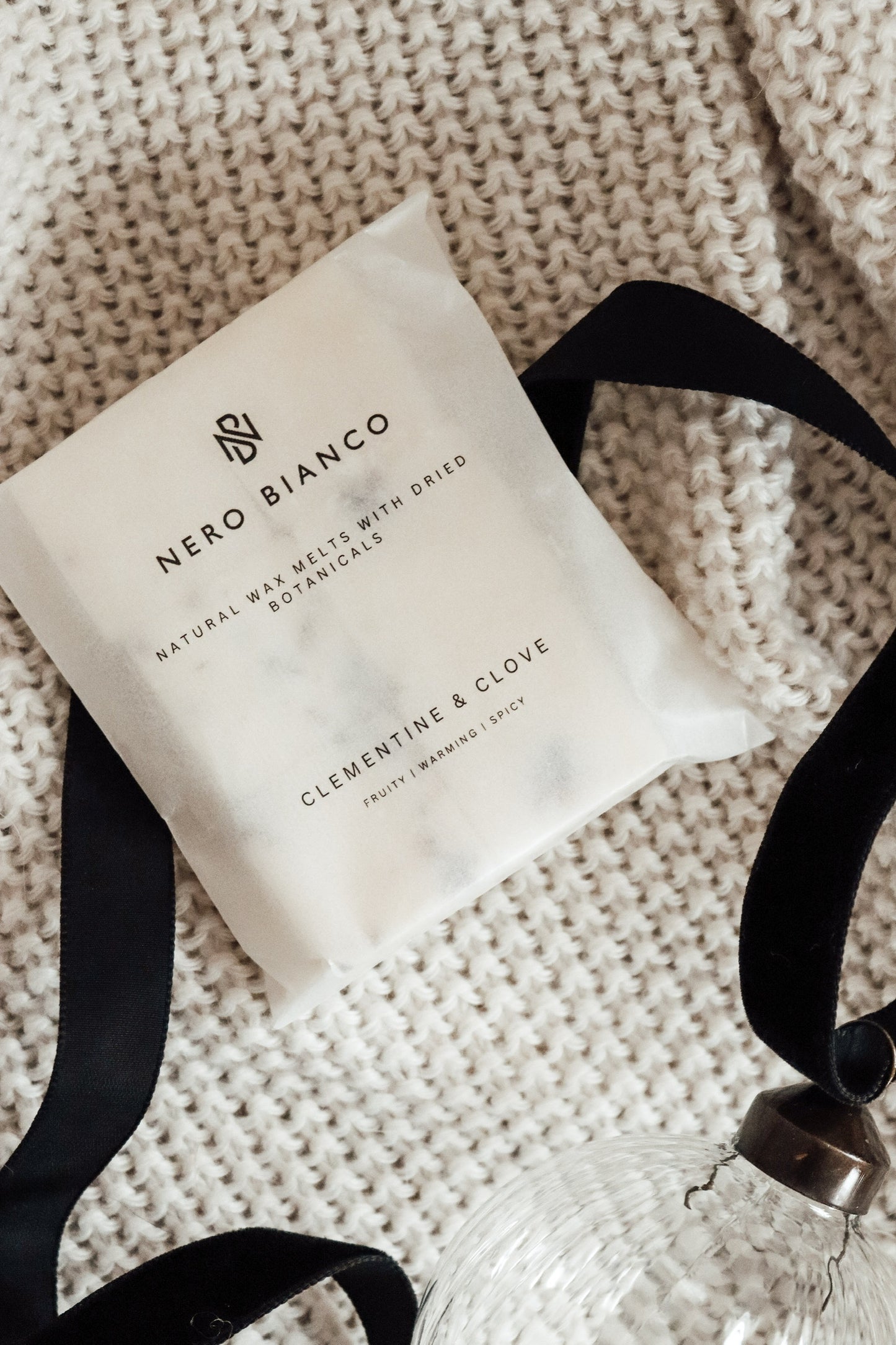 scented wax melts in a glassine pack lying on a beige blanket with black ribbon