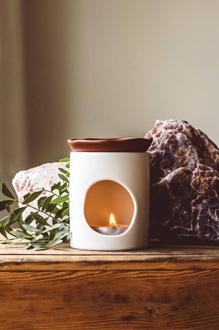 White wax melt burner with a small candle inside on a wooden surface with rocks and leaves.