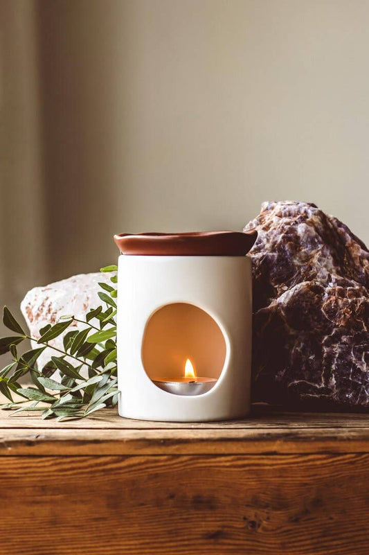 White wax melt burner with a small candle inside on a wooden surface with rocks and leaves.