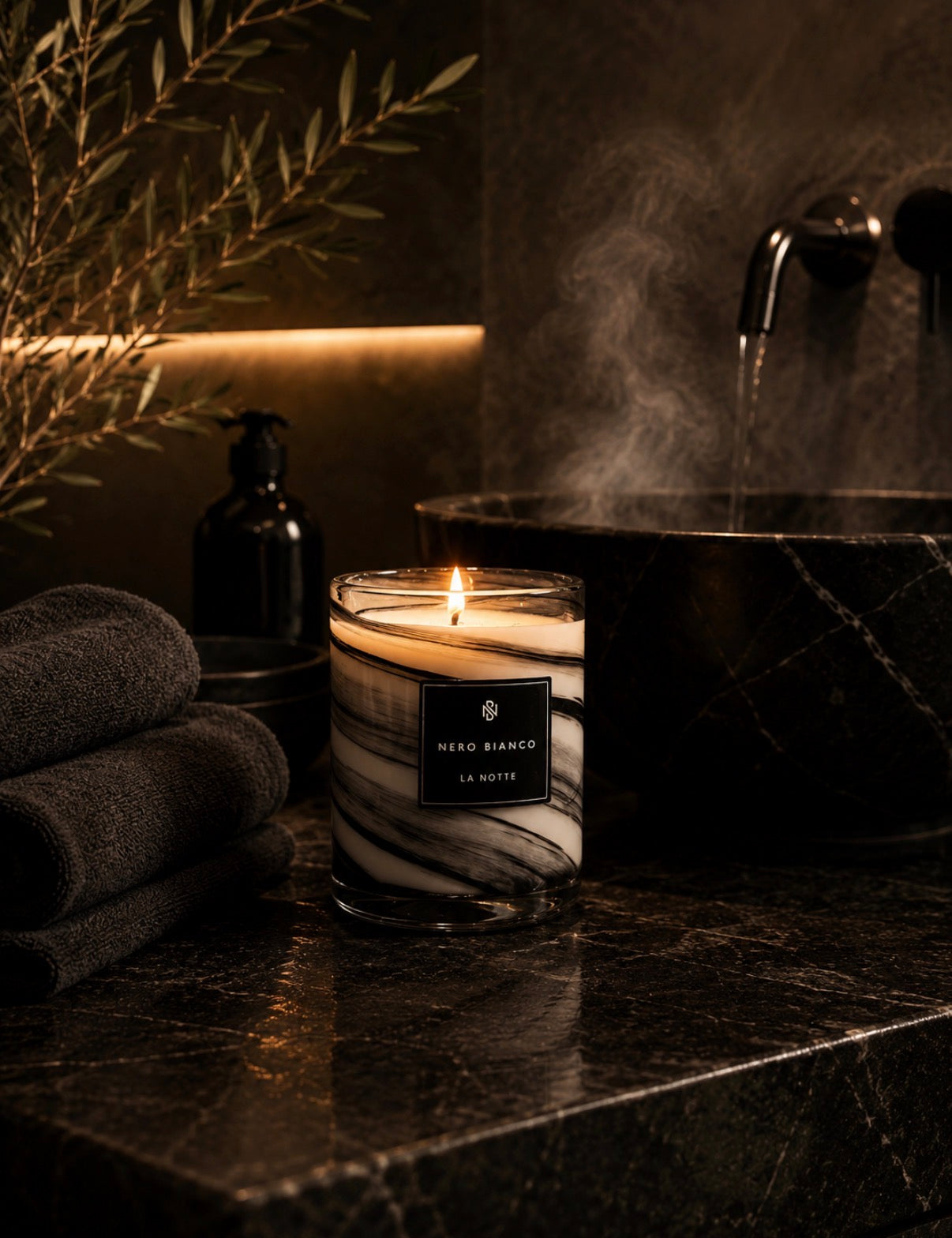 A lit scented candle in a clear glass jar with a black and white marble pattern, placed on a dark marble bathroom counter next to a steaming sink, folded towels, and a soap dispenser. The label reads 'NEODIOXIDE LA NOTTE'.