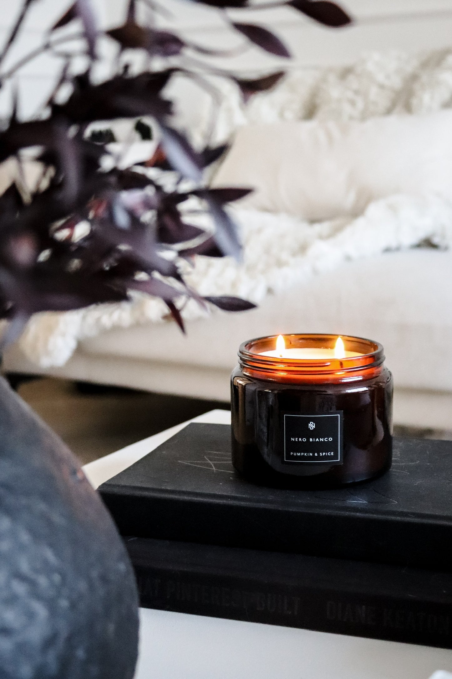 An amber glass candle with a black lid, placed on a table with a plant and a book in the background, with a cozy room setting.