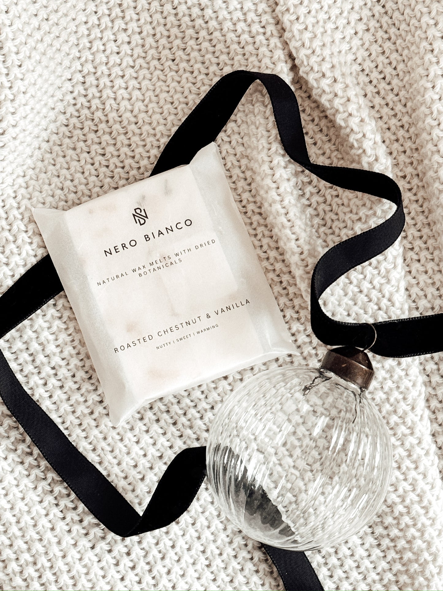 wax melts in a glassine pack lying on a beige blanket with a glass bauble and black ribbon