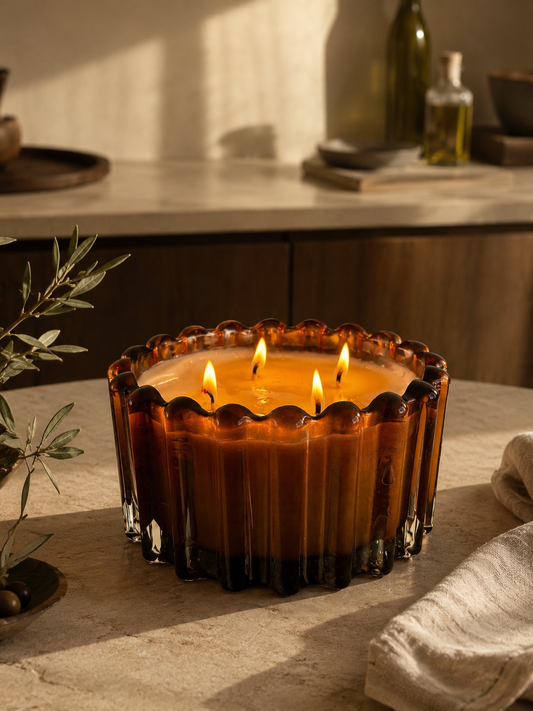 A large amber glass candle with three lit wicks sits on a stone countertop in a warm, rustic kitchen setting.