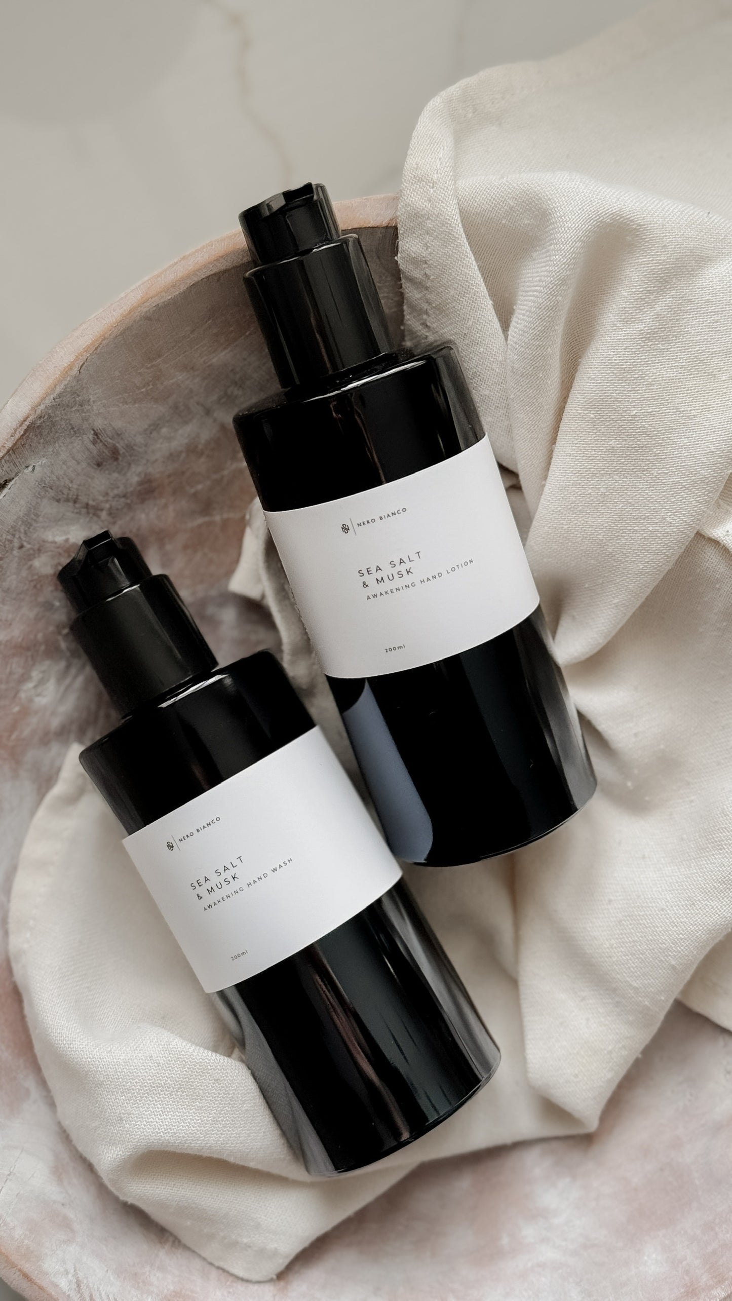Two black skincare bottles with white labels on a beige fabric background