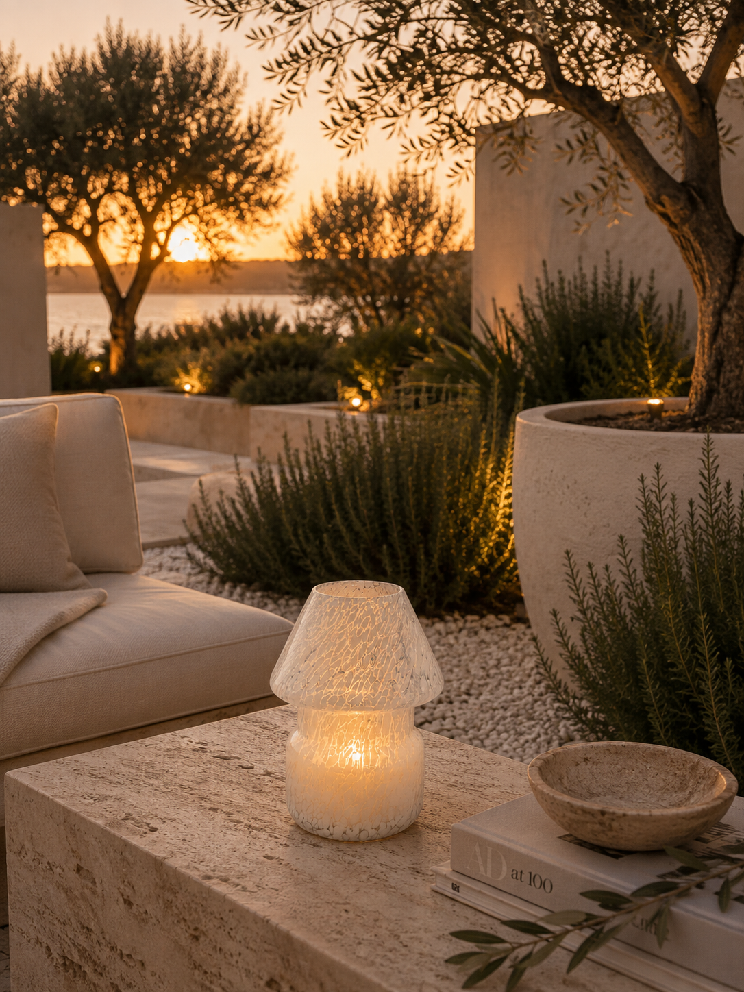 A lit textured glass candle holder with a mushroom-shaped shade sits on a stone table in an outdoor patio setting at sunset, surrounded by plants, a sofa, and warm ambient lighting.