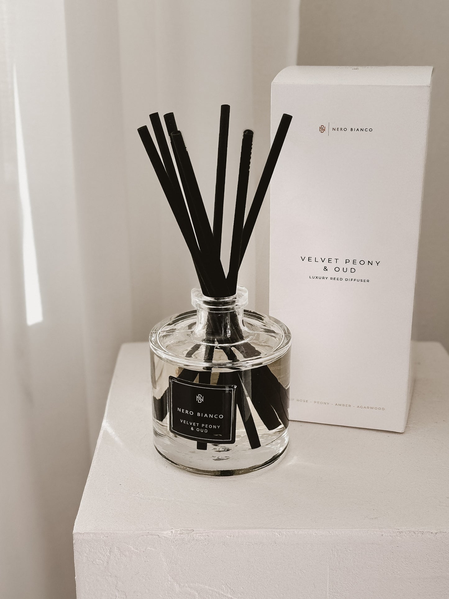 Reed diffuser with black reeds and a white box labeled 'Velvet Peony & Oud' on a white surface