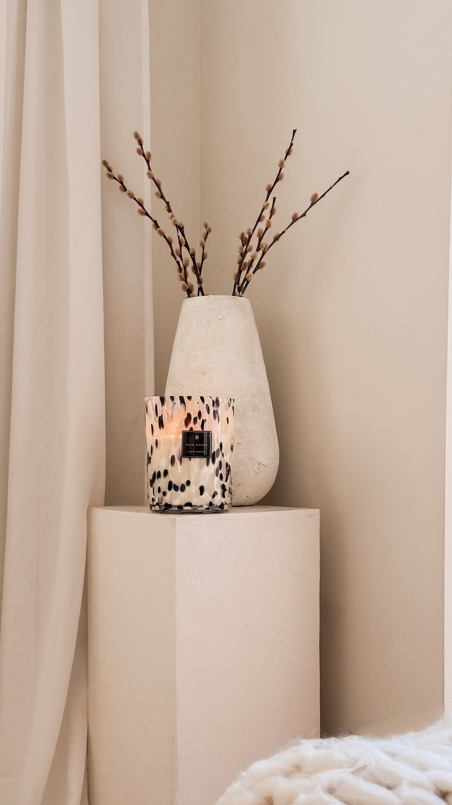 Candle in a decorative vase with branches on a beige pedestal against a beige curtain.