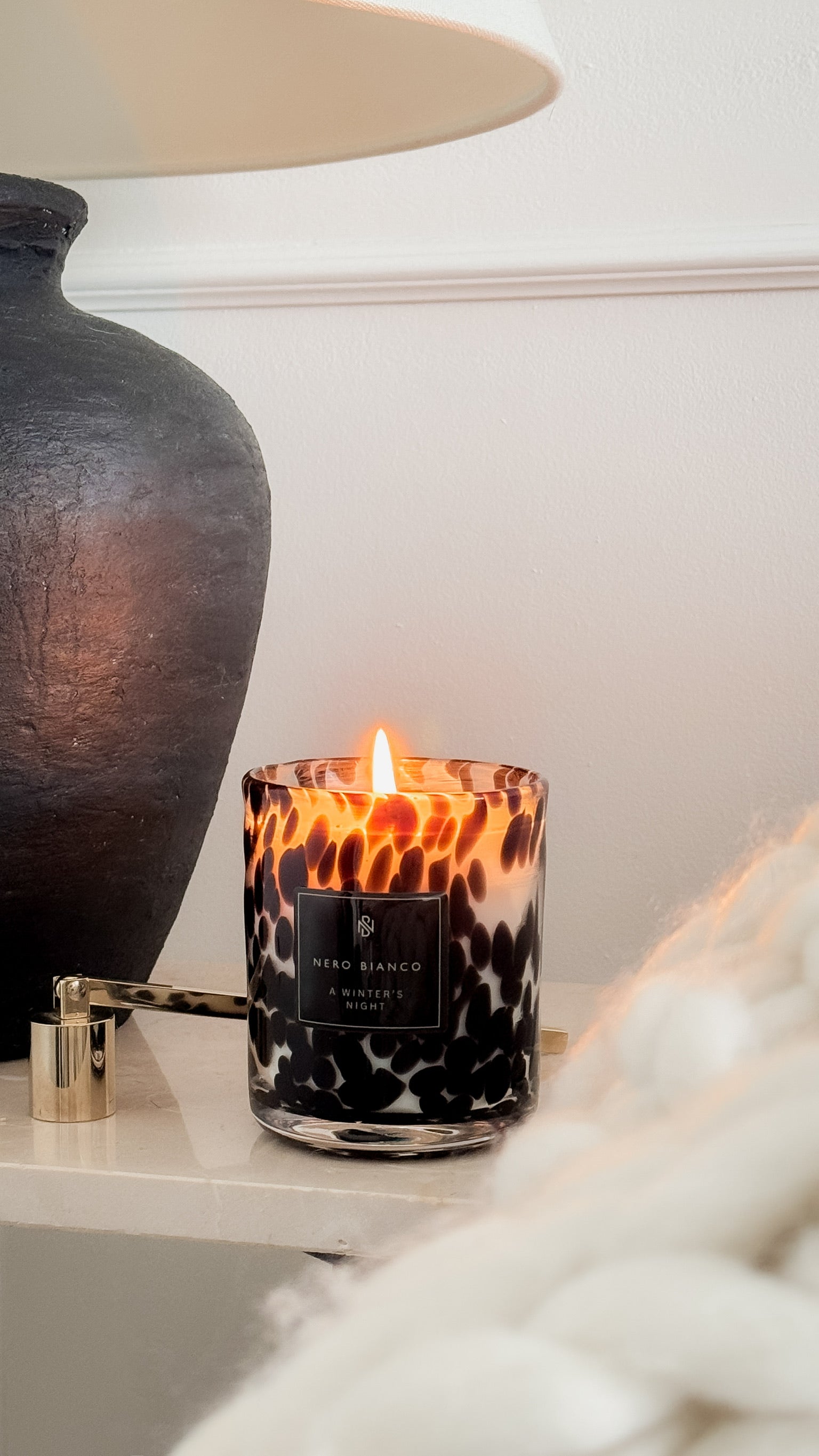 Candle with a black and white pattern on a surface next to a lamp and decorative vase.