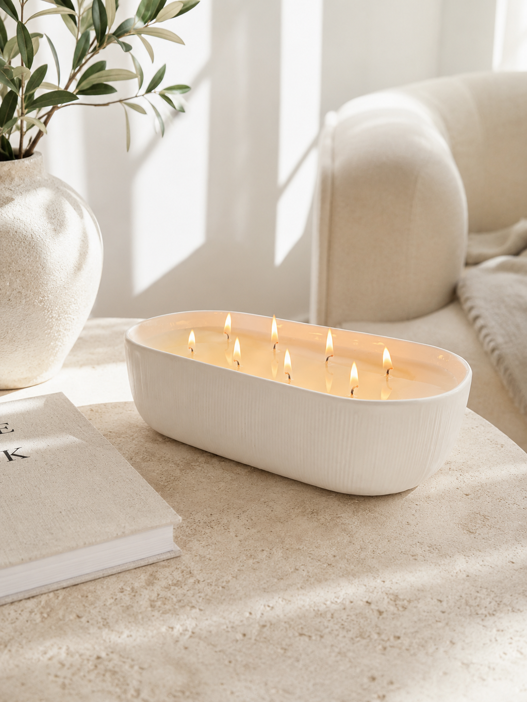 A white oval candle holder with multiple lit wicks, placed on a light stone table in a softly lit room with a sofa, book, and vase in the background.