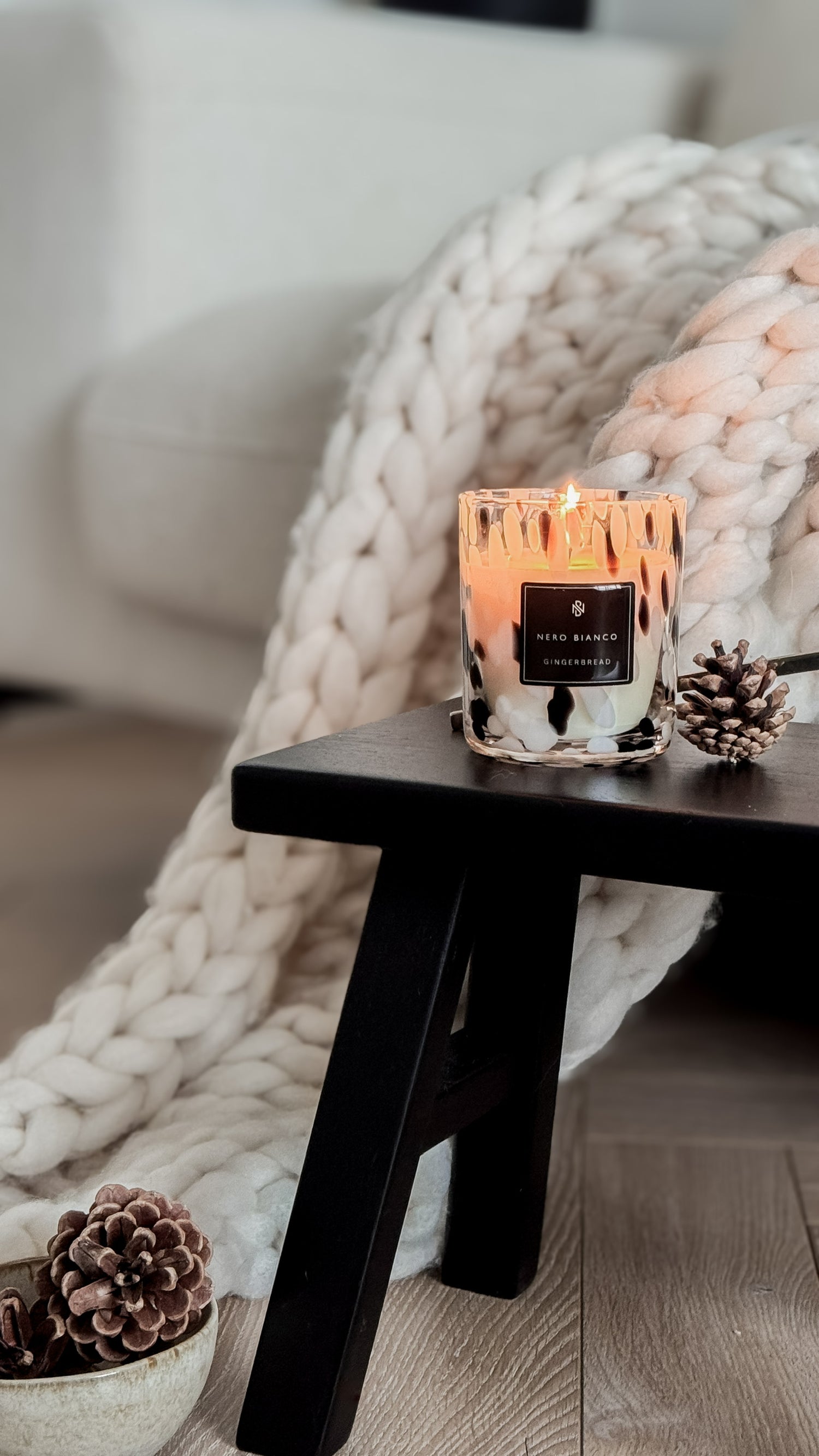 Candle on a small wooden table with a cozy blanket and pinecones in the background