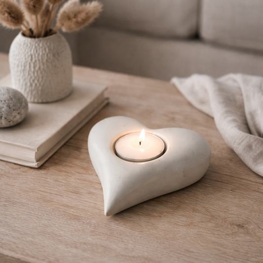 Heart-shaped candle holder with a lit candle on a wooden surface, with a vase and books in the background.