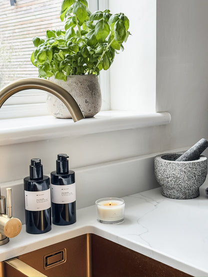 Candle on a kitchen counter with luxury hand wash and lotion in black bottles and a potted basil in the background