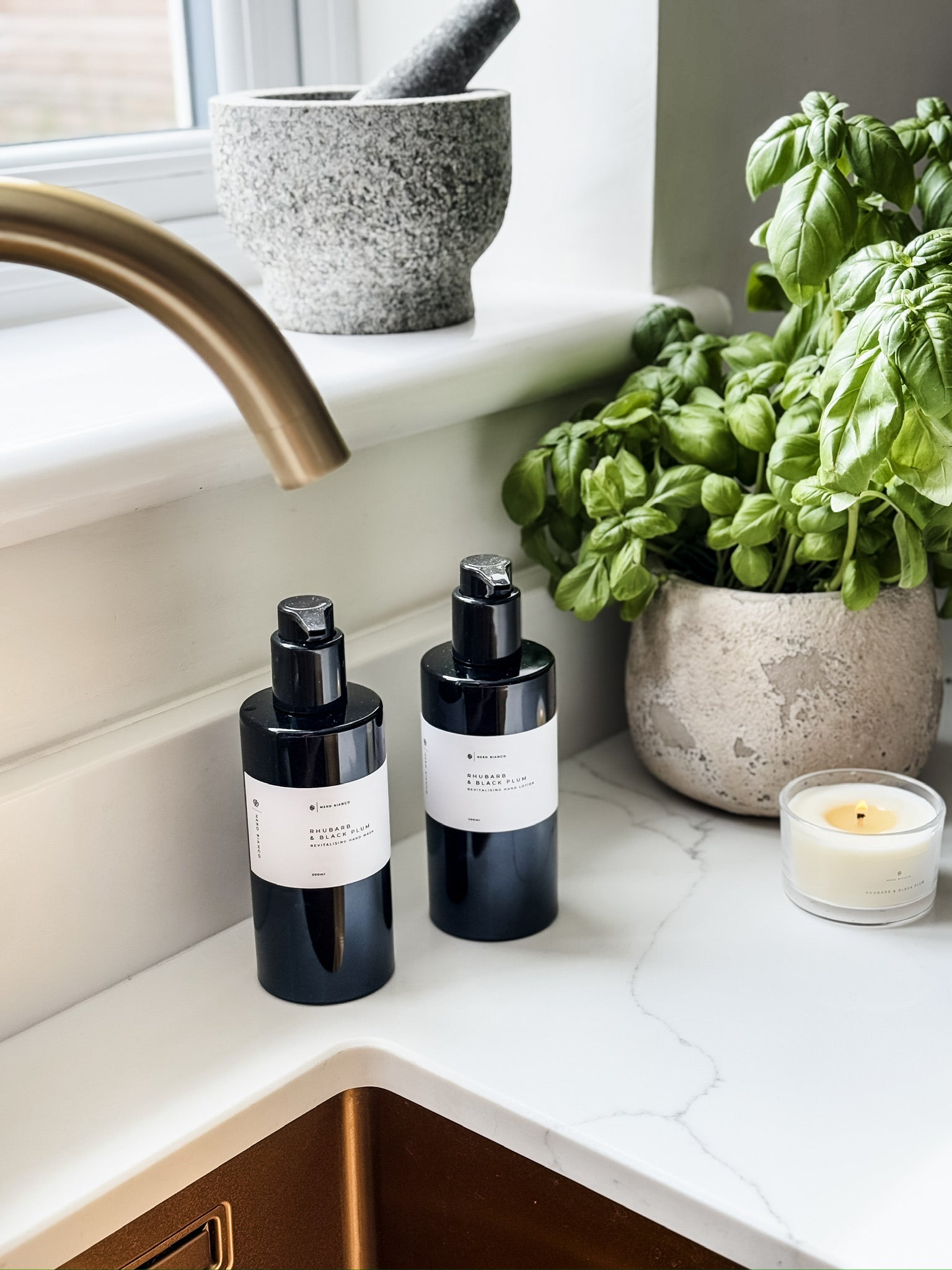 hand wash and hand lotion in black bottles with white labels on a kitchen counter next to a basil plant and candle.
