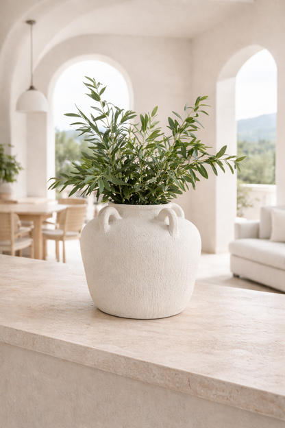 White vase with green plant on a wooden surface in a bright room with arched windows.