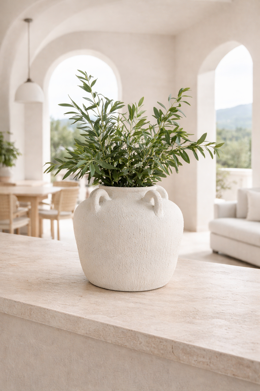 White vase with green plant on a wooden surface in a bright room with arched windows.