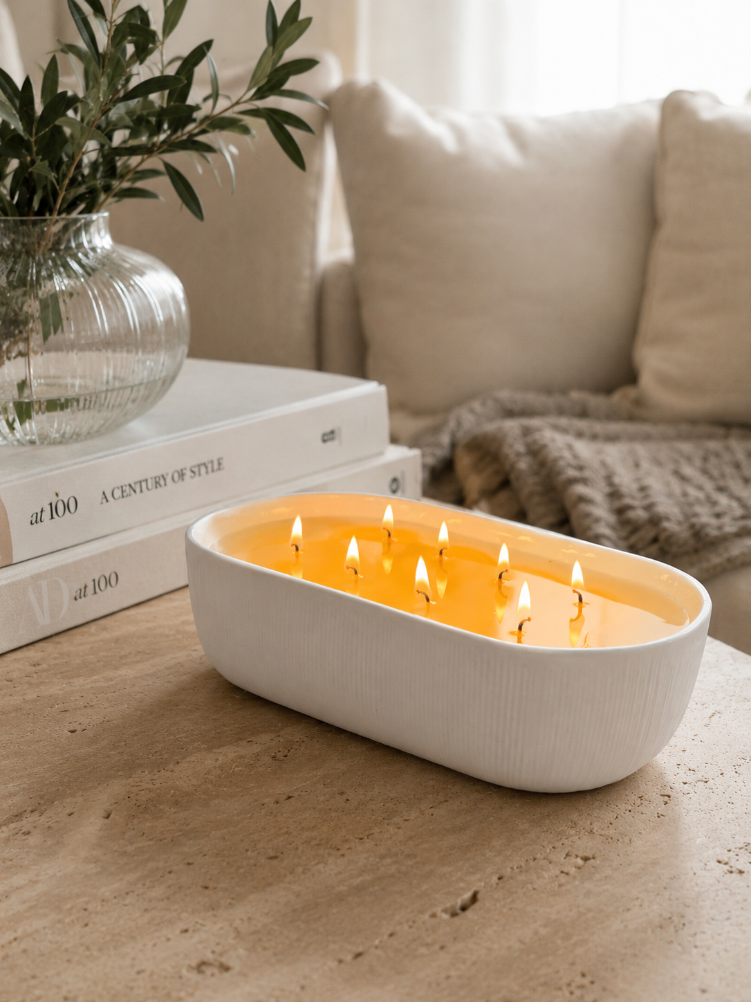 A white oval candle holder filled with multiple lit wicks, placed on a stone surface in a cozy living room setting with books and a vase of greenery in the background.