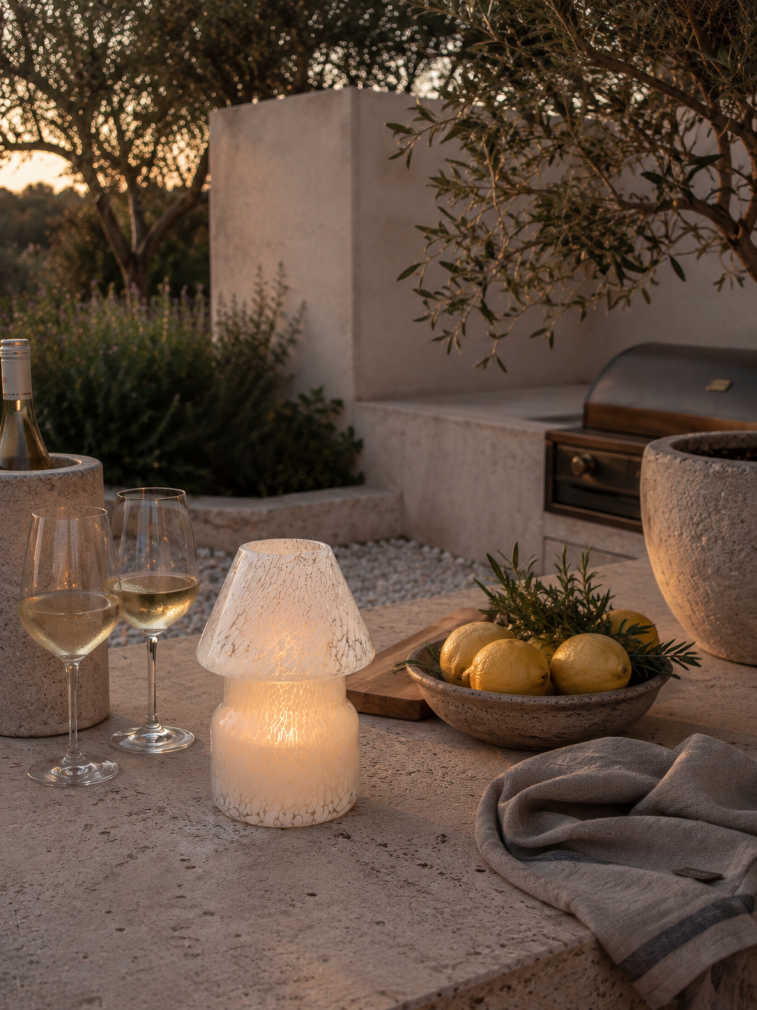 A glowing mushroom-shaped candle holder with a textured white glass body and a matching textured white lampshade, styled on a stone patio alongside wine glasses, lemons, and a stone basket in an outdoor evening setting.