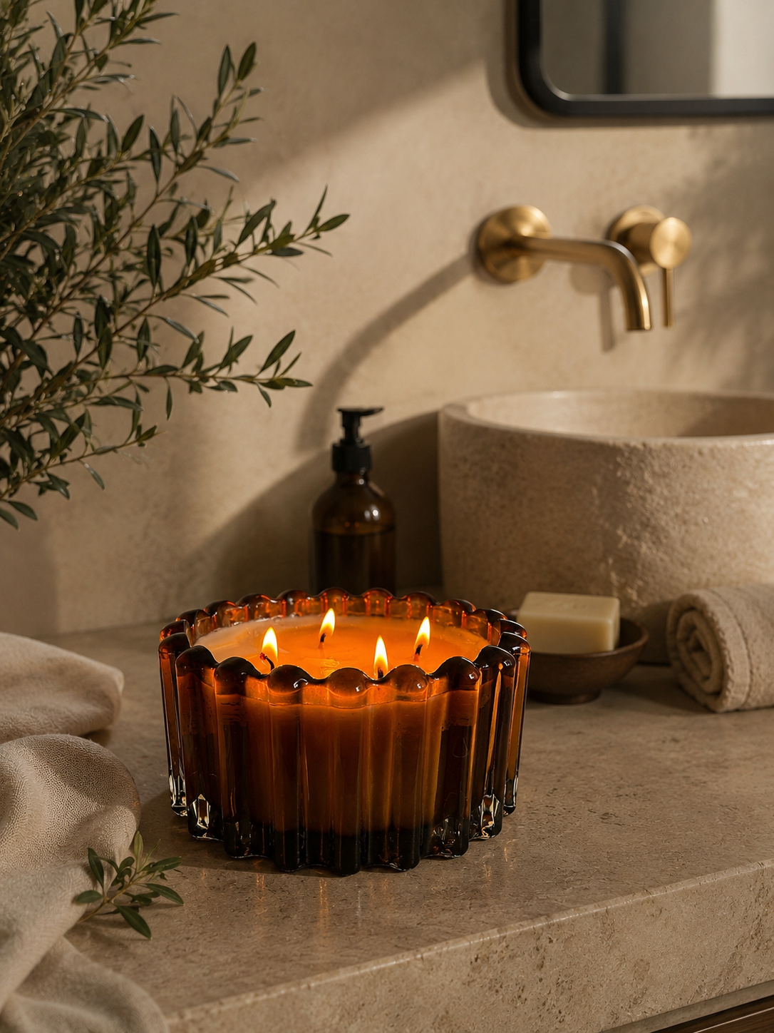 A lit amber glass candle holder with multiple flames sits on a stone bathroom counter beside a sink, soap dish, soap bar, towel, and a dark bottle, with warm wall lighting and a mirror in the background.