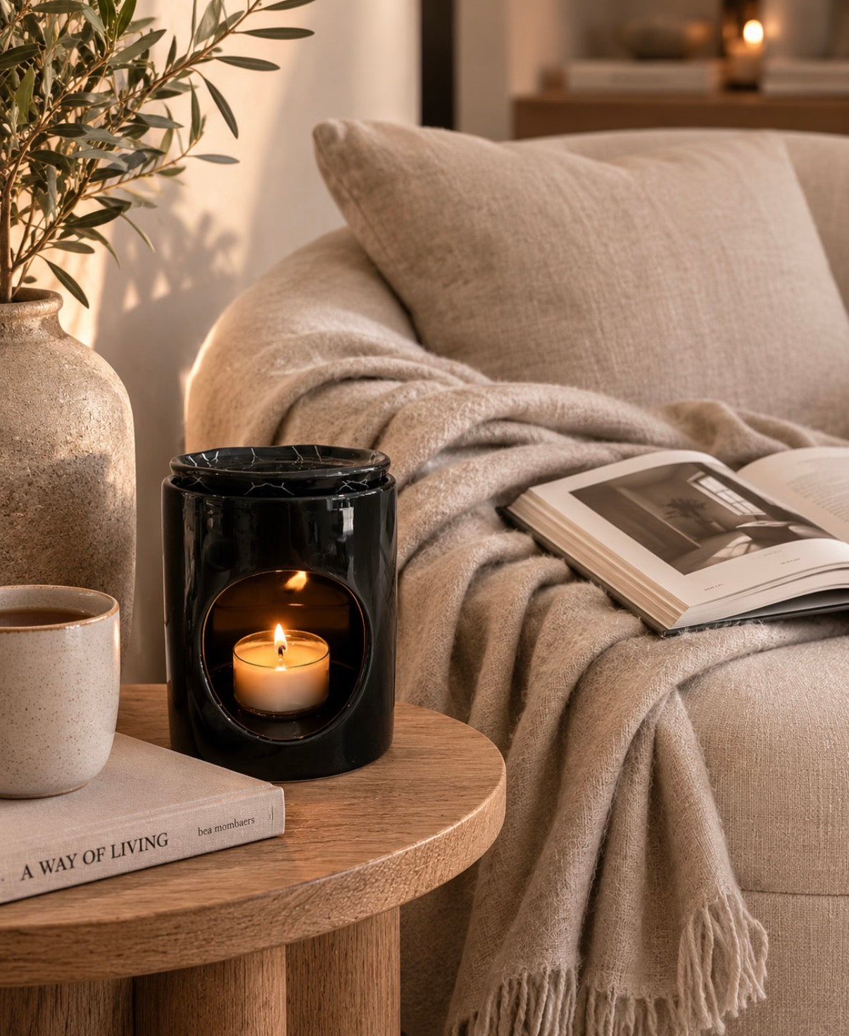 A black glass candle holder with a lit candle inside, styled on a round wooden side table next to a mug, books, and a cozy beige throw blanket draped over a sofa in a warm living room setting.