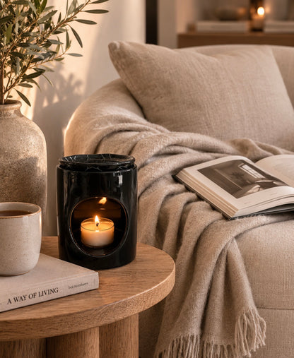 A black glass candle holder with a lit candle inside, styled on a round wooden side table next to a mug, books, and a cozy beige throw blanket draped over a sofa in a warm living room setting.