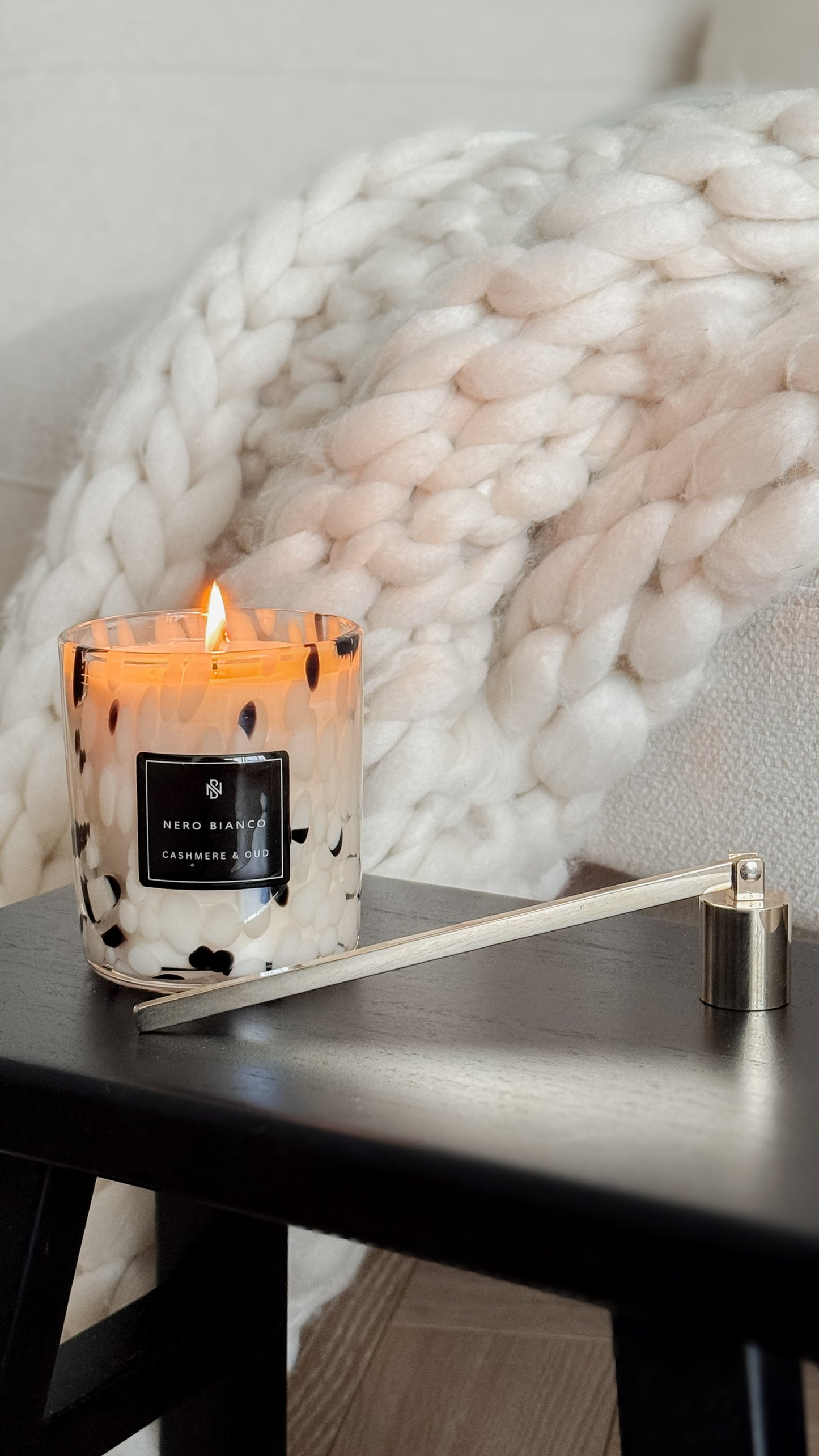 Candle with a lit wick on a wooden surface in front of a textured white wall.