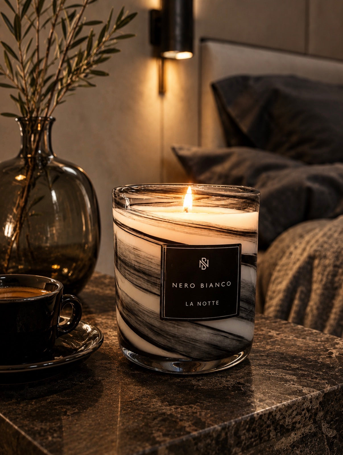 A lit candle in a clear glass jar with a black and white marbled label reading 'NERO BIANCO LA NOTTE' sits on a dark stone bedside table in a cozy bedroom setting. A cup of coffee, a glass vase with dried stems, and a bed are visible in the background.