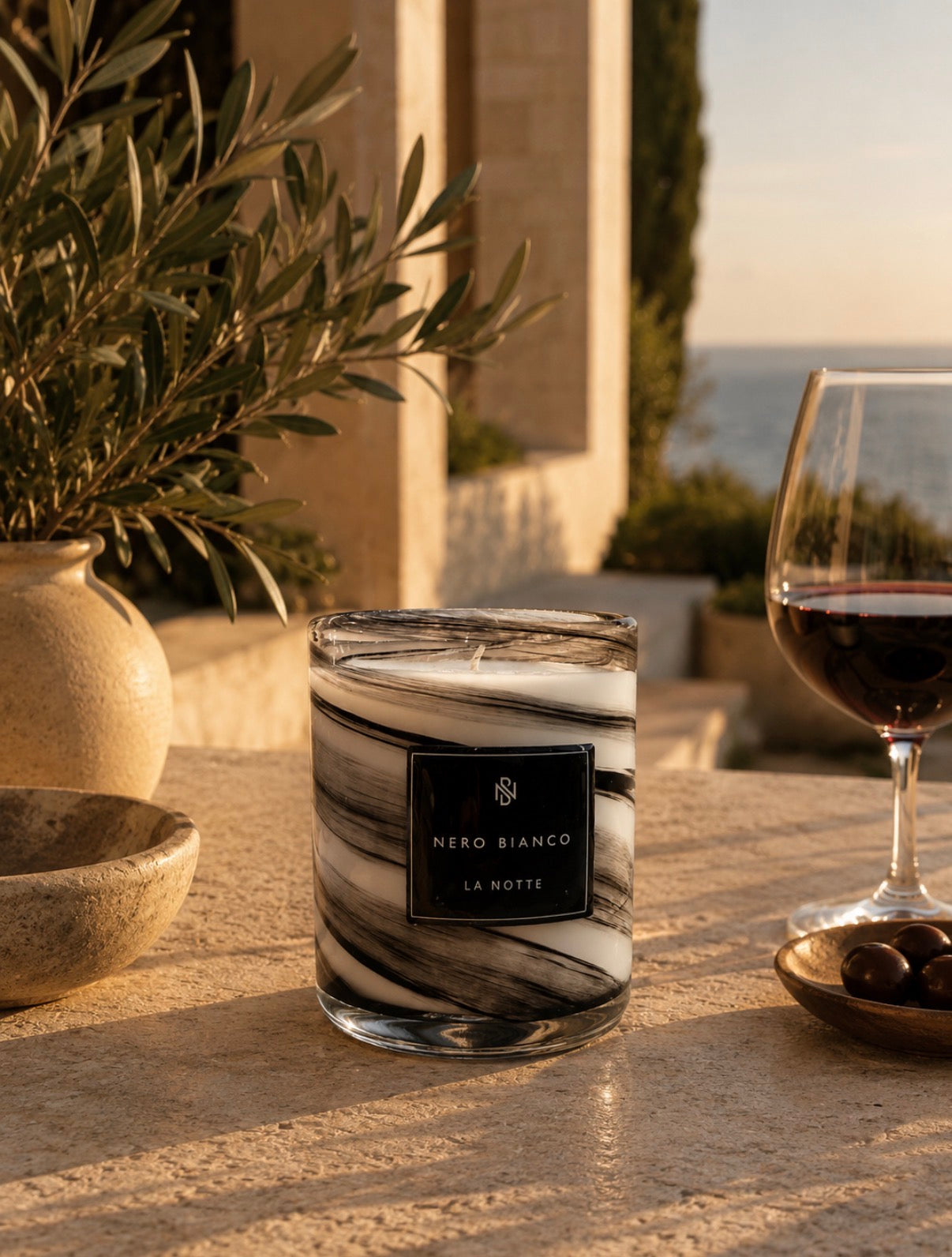 A scented candle in a clear glass jar with black and white marble swirls sits on a stone table outdoors. A wine glass with red wine and a potted olive tree are blurred in the background, with the sea and sky visible behind them.