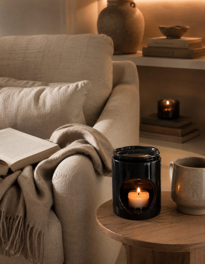 A black glass candle holder with a lit candle sits on a wooden side table next to a beige armchair with a throw blanket and an open book in a cozy, warmly lit living room.