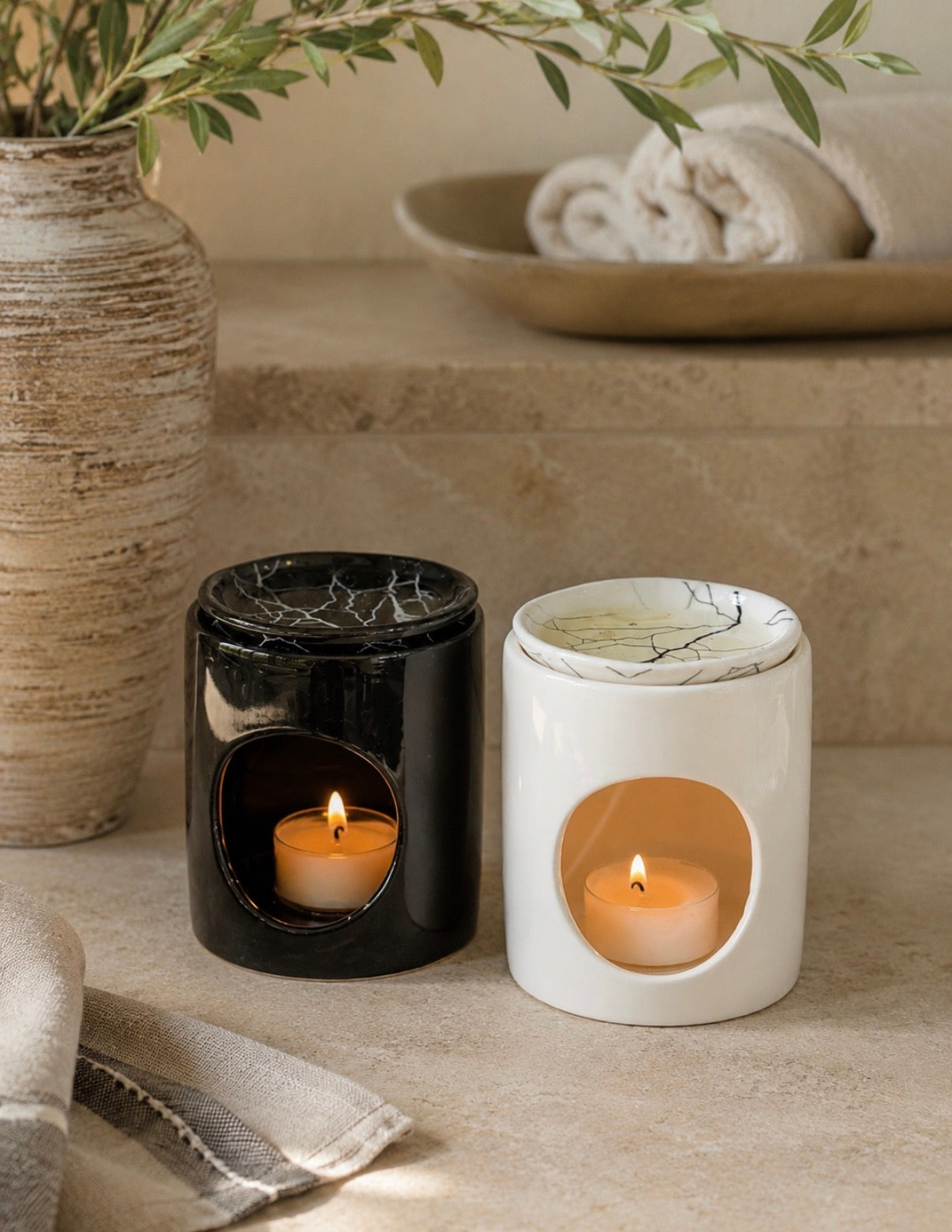 Two ceramic candle holders, one glossy black and one matte white with a marble-look pattern, each containing a lit tea light candle. They are styled on a beige stone surface next to a textured vase with greenery and a folded towel.