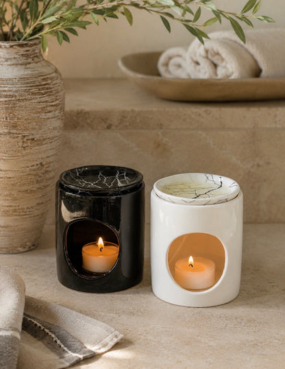 Two ceramic candle holders, one glossy black and one matte white with a marble-look pattern, each containing a lit tea light candle. They are styled on a beige stone surface next to a textured vase with greenery and a folded towel.