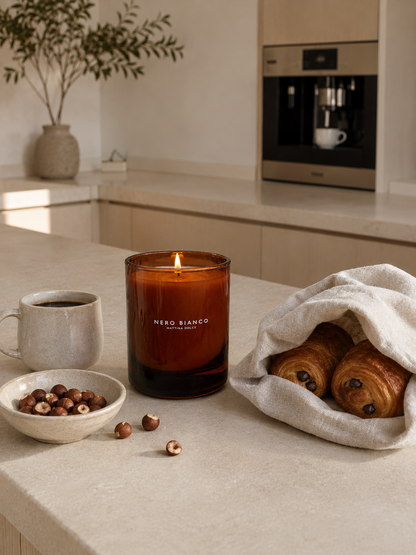 A lit amber glass scented candle with the brand name 'NERO BIANCO' on the front, styled on a beige countertop with a coffee mug, a bowl of hazelnuts, and pastries in a warm kitchen setting.
