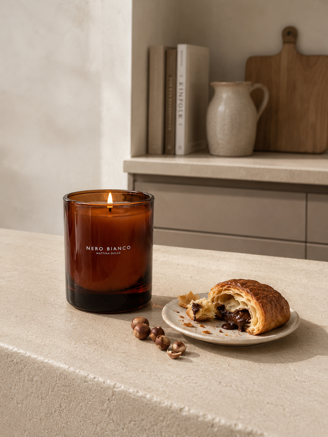 An amber glass scented candle by NERO BIANCO sits lit on a beige stone countertop beside a partially eaten pastry on a small plate with a few hazelnuts. A kitchen shelf with books, a ceramic jug, and a wooden cutting board is softly blurred in the background.