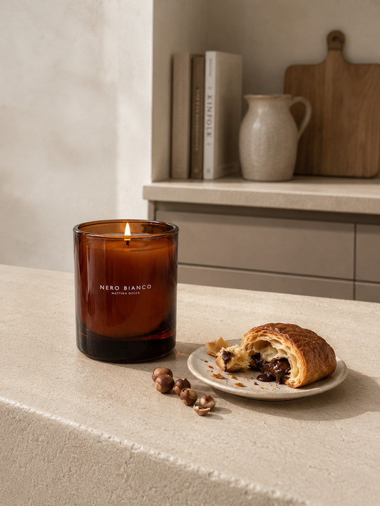 An amber glass scented candle by NERO BIANCO sits lit on a beige stone countertop beside a partially eaten pastry on a small plate with a few hazelnuts. A kitchen shelf with books, a ceramic jug, and a wooden cutting board is softly blurred in the background.