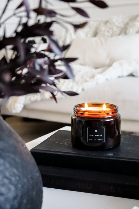 An amber glass candle with a black lid, placed on a table with a plant and a book in the background, with a cozy room setting.