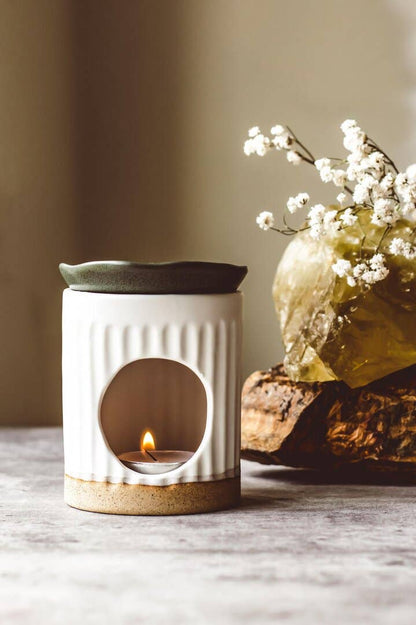 White ceramic diffuser with a small flame and a vase of white flowers on a wooden surface.