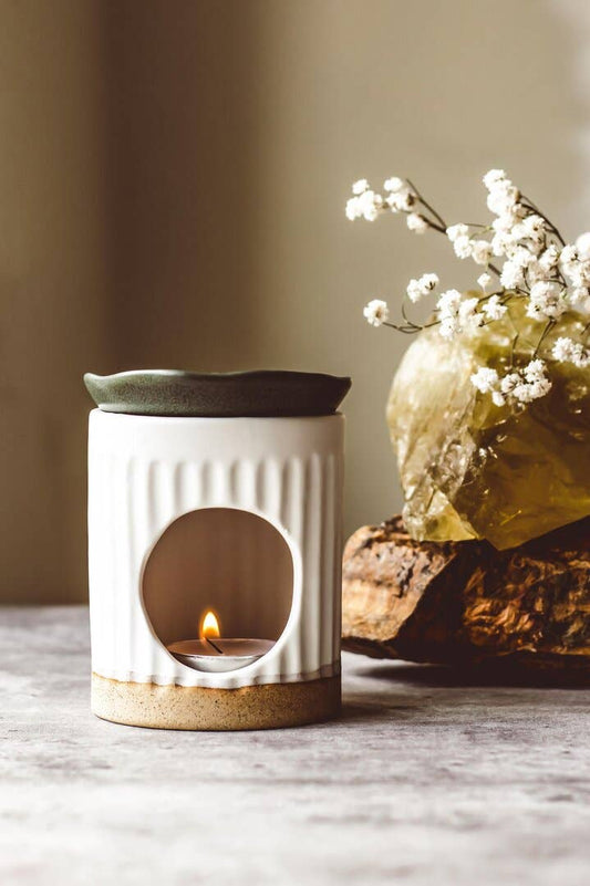 White ceramic diffuser with a small flame and a vase of white flowers on a wooden surface.