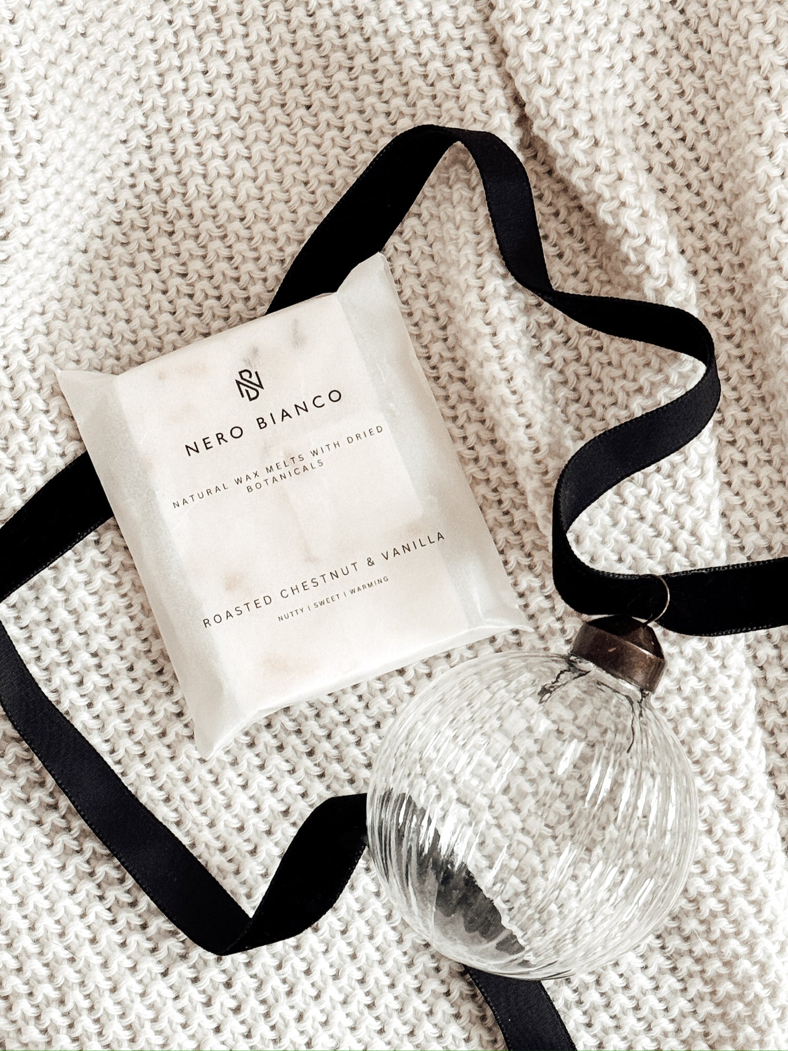 wax melts in a glassine pack lying on a beige blanket with a glass bauble and black ribbon