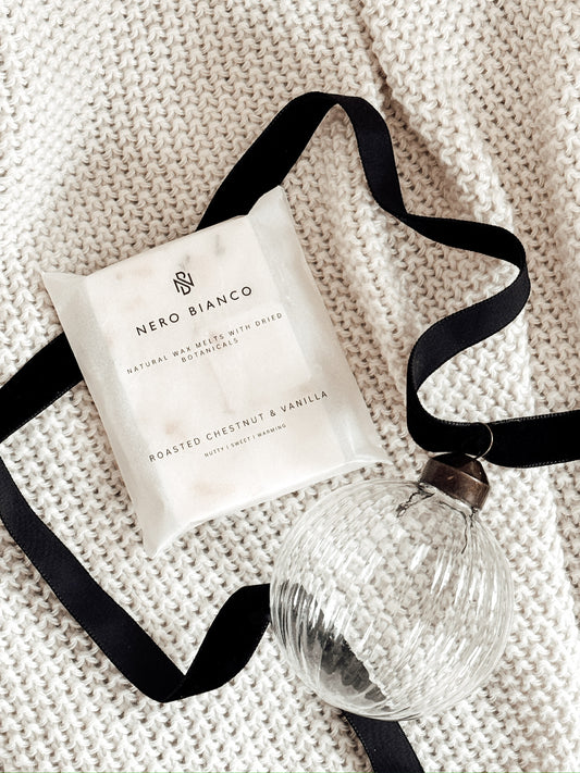 wax melts in a glassine pack lying on a beige blanket with a glass bauble and black ribbon