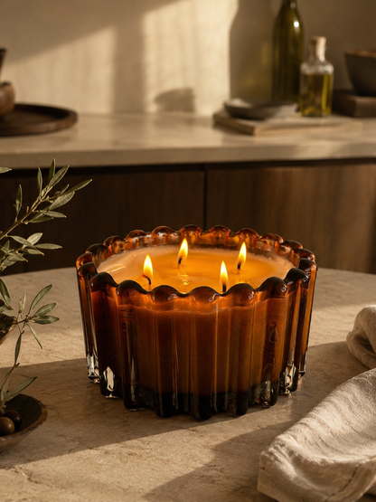A large amber glass candle with three lit wicks sits on a stone countertop in a warm, rustic kitchen setting.