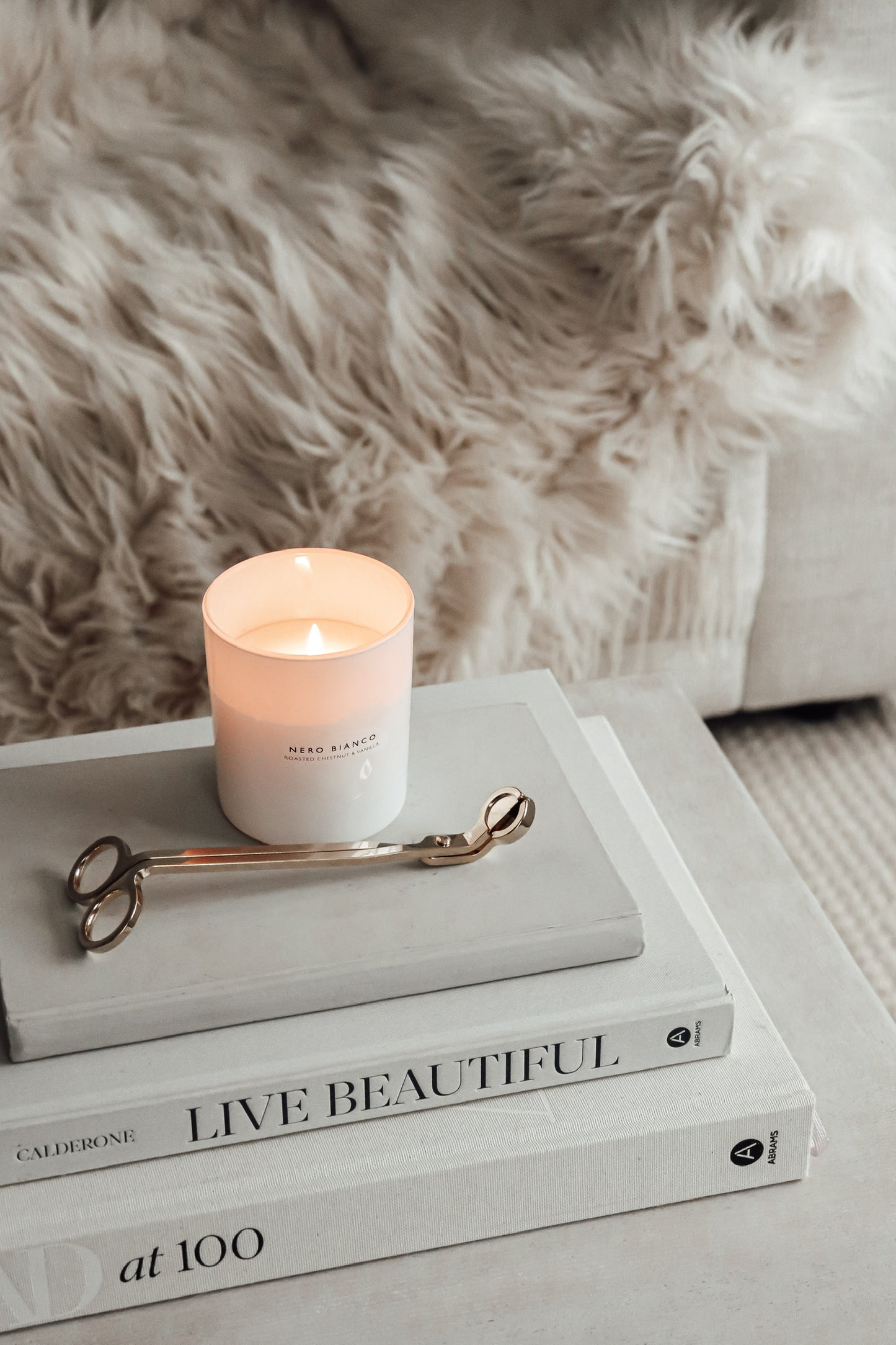 A lit white candle with on top of a stack of books with the titles. The candle is placed next to gold candle wick snippers on a faux fur surface.