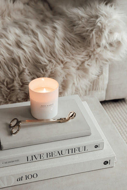 A lit white candle with on top of a stack of books with the titles. The candle is placed next to gold candle wick snippers on a faux fur surface.