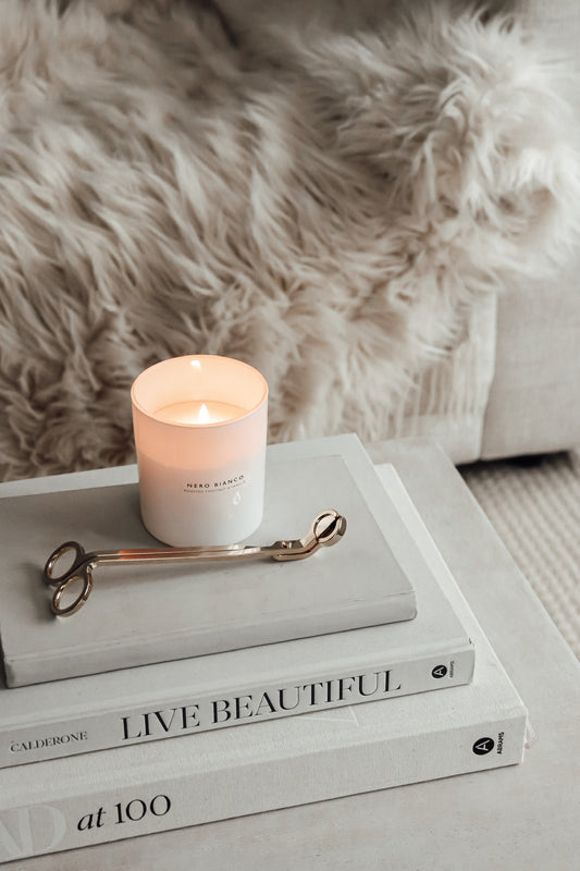 A lit white candle with on top of a stack of books with the titles. The candle is placed next to gold candle wick snippers on a faux fur surface.
