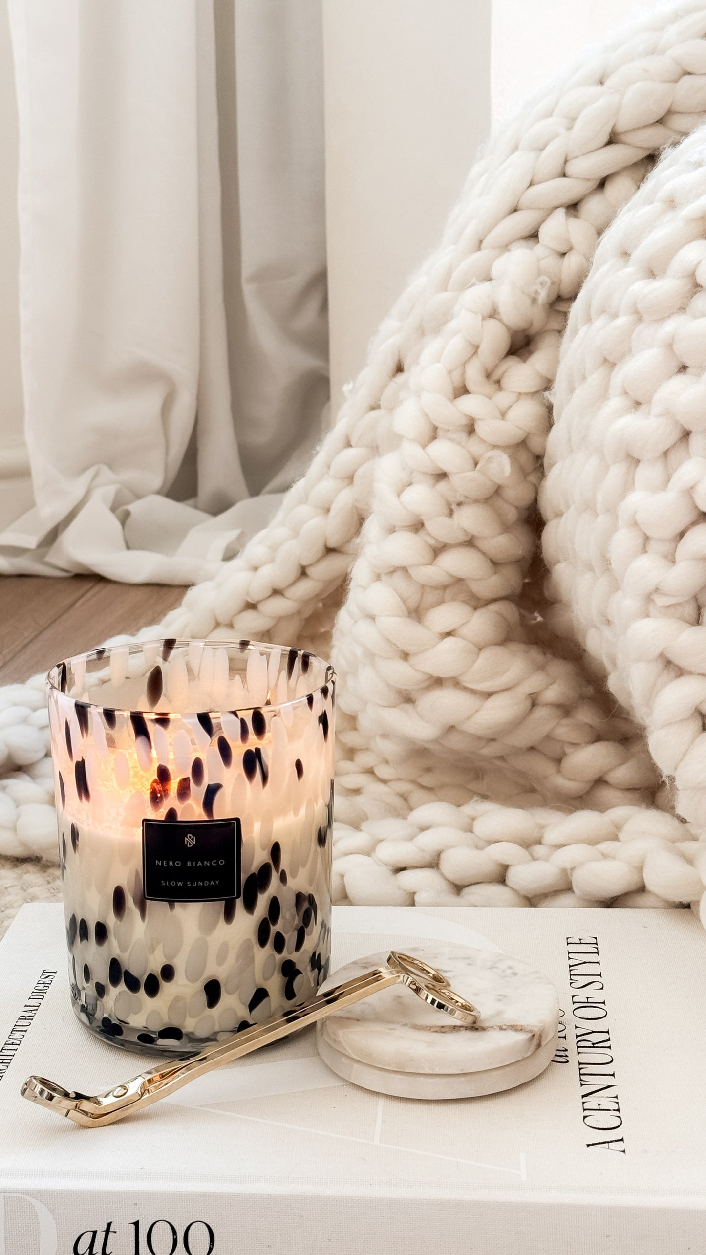 Candle with decorative packaging on a surface with a blanket and magazine in the background