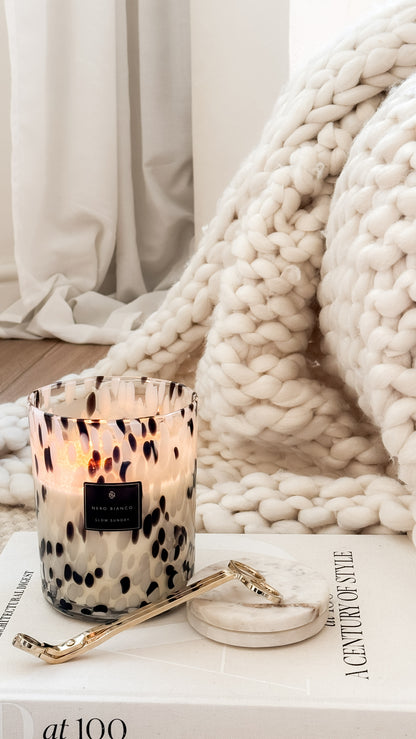 Candle with decorative packaging on a surface with a blanket and magazine in the background