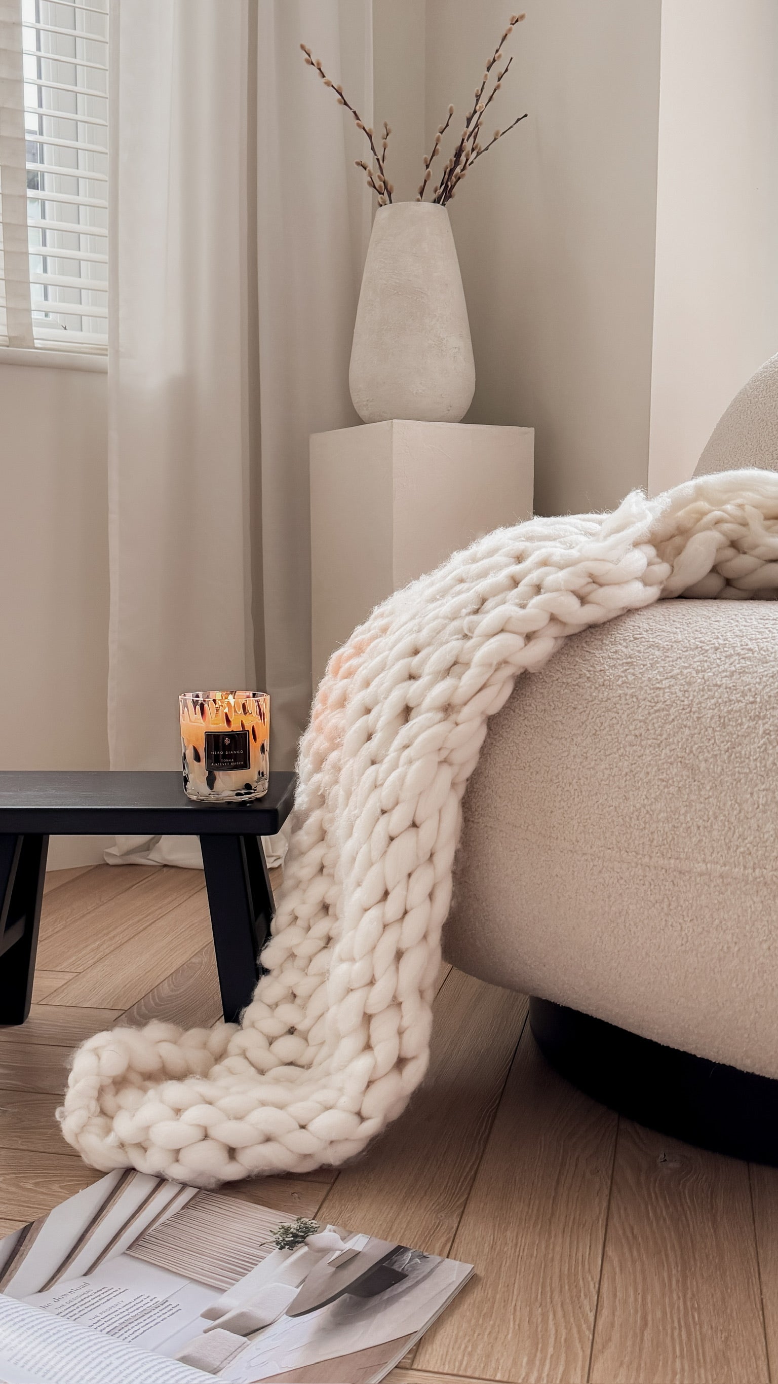 Cozy living room with a white knit blanket draped over a sofa, a candle on a small table, and a vase with branches.
