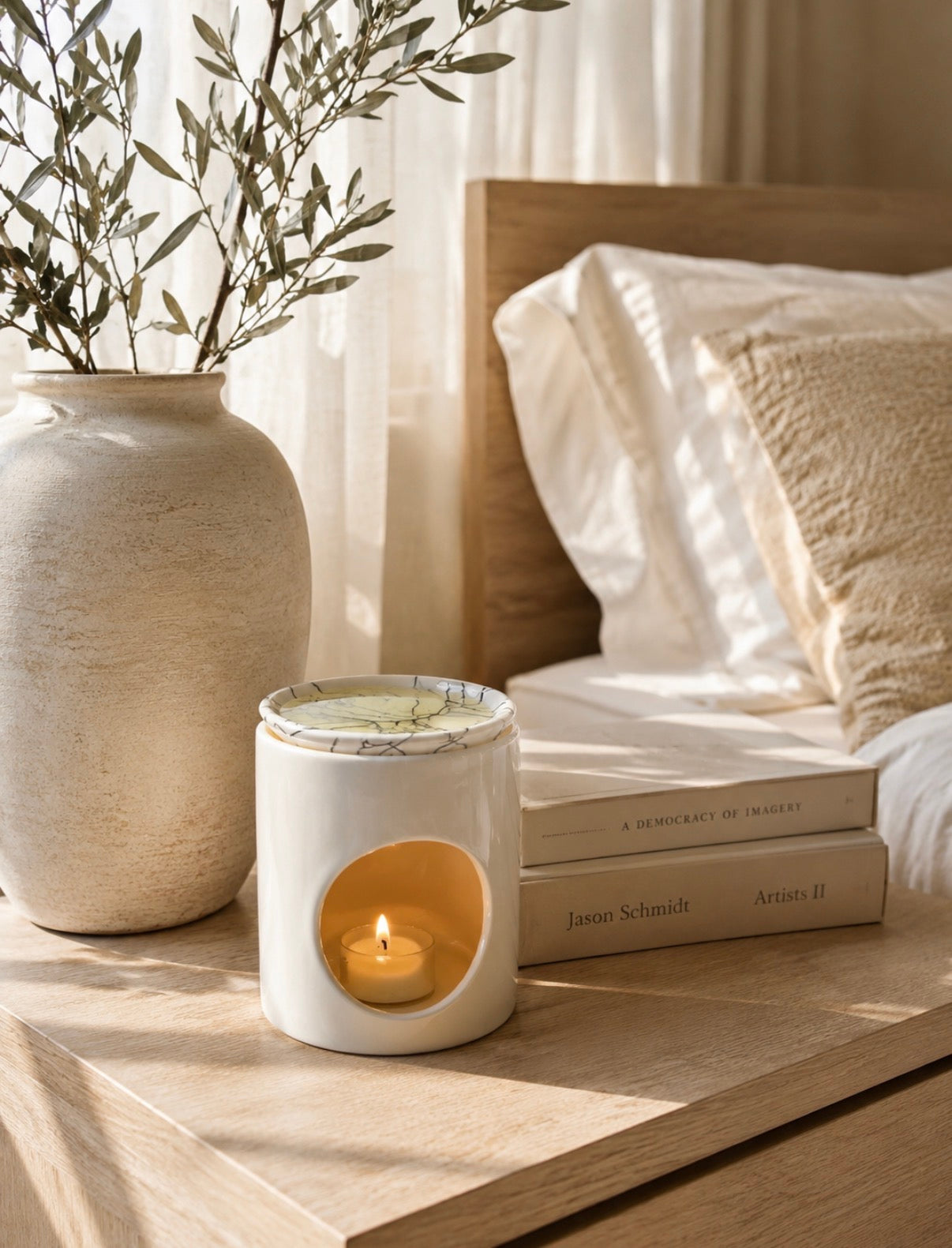 A white ceramic candle holder with a lit tea light candle sits on a light wood bedside table next to a large textured vase with olive branches and stacked books in a softly lit bedroom setting.